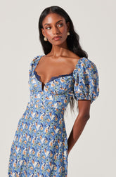 A woman is wearing a dress with a blue floral pattern that features orange, white, and green accents. The dress has puffed short sleeves and a sweetheart neckline edged with dark lace trim.